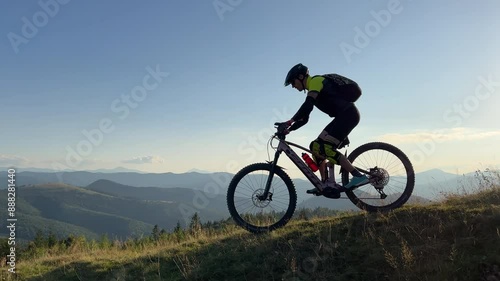 Cyclist man riding electric mountain bike outdoors. Silhouette of male tourist biking along grassy trail in the mountains, wearing helmet and backpack. Concept of sport, active leisure and nature.