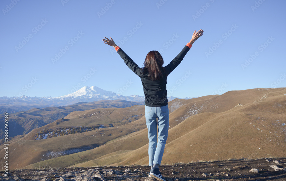 A young brunette woman with her hands raised up against the backdrop of the snow-capped Mount Elbrus.