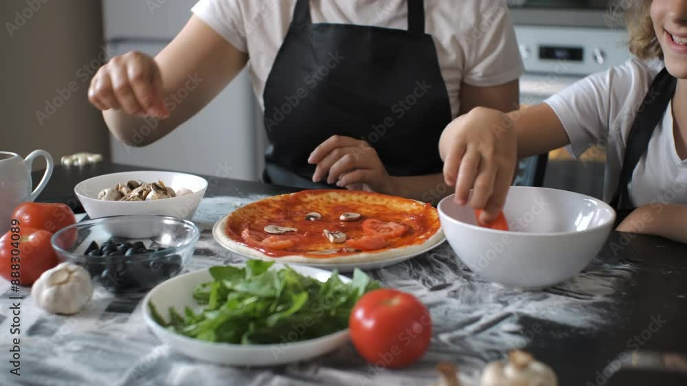 In cozy kitchen, a mother is placing slices of mushrooms onto a pizza crust topped with sauce, while her daughter adds fresh tomato slices. Slow motion. Family collaboration and healthy cooking In cozy kitchen, a mother is placing slices of mushrooms onto a pizza crust topped with sauce, while her daughter adds fresh tomato slices. Slow motion. Family collaboration and healthy cooking