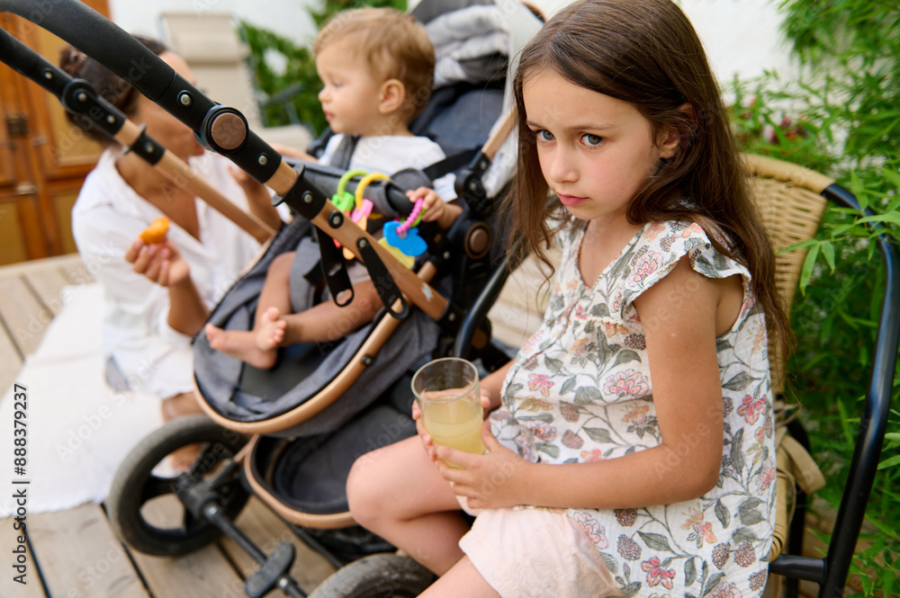 Obraz premium Young girl holding a glass of juice while sitting next to a baby in a stroller and mother. Scene captures outdoor family time and sibling interaction.