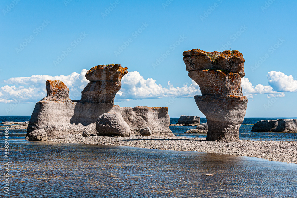 Fototapeta premium Breathtaking limestone sculptures rise from a shallow coastal pool at Mingan Archipelago National Park Reserve, framed by a clear blue sky adorned with dramatic clouds.