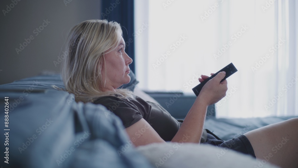 Side view of woman watching television or TV shows while sitting on comfortable sofa and uses remote control to change TV content. Caucasian woman relaxing at home in living room. Lifestyle concept. Side view of woman watching television or TV shows while sitting on comfortable sofa and uses remote control to change TV content. Caucasian woman relaxing at home in living room. Lifestyle concept.