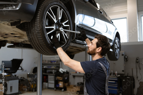 Male mechanic fixing tire of elevated car in modern auto repair shop. Professional auto service technician working with tools in garage. Vehicle maintenance and repair concept.