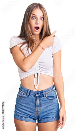 Young beautiful blonde woman wearing casual white tshirt surprised pointing with finger to the side, open mouth amazed expression.