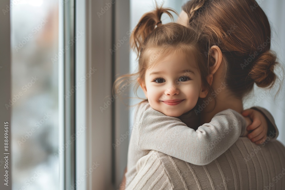 Cute little girl with pigtails hugging her mom, both smiling, standing by a large window with soft daylight, minimalistic background, waist-length photo. Cute little girl with pigtails hugging her mom, both smiling, standing by a large window with soft daylight, minimalistic background, waist-length photo.