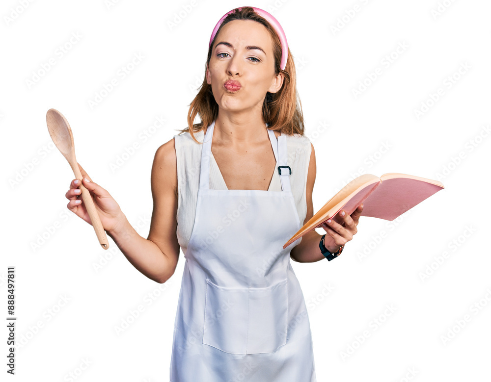 Young caucasian woman wearing professional baker apron reading cooking recipe book looking at the camera blowing a kiss being lovely and sexy. love expression. Young caucasian woman wearing professional baker apron reading cooking recipe book looking at the camera blowing a kiss being lovely and sexy. love expression.