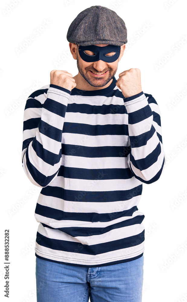 Young handsome man wearing burglar mask excited for success with arms raised and eyes closed celebrating victory smiling. winner concept. Young handsome man wearing burglar mask excited for success with arms raised and eyes closed celebrating victory smiling. winner concept.