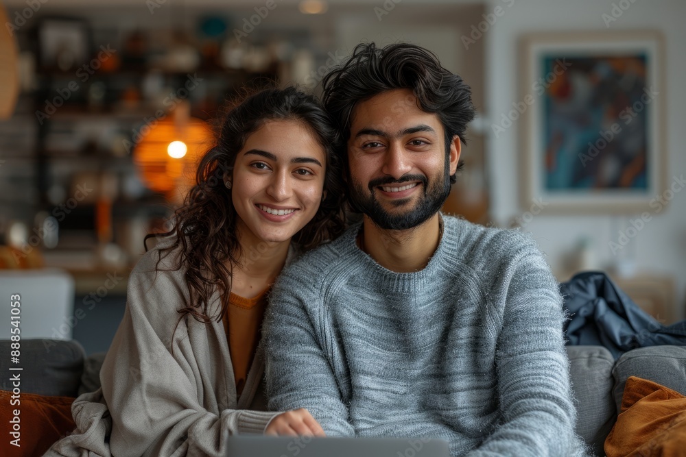 A smiling young Indian couple sit on a gray sofa in a modern living room at home. They use a laptop computer together to make online shopping or have a video call with family during their free time, i A smiling young Indian couple sit on a gray sofa in a modern living room at home. They use a laptop computer together to make online shopping or have a video call with family during their free time, i