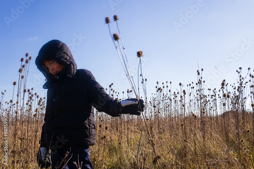 A boy in a black hooded down jacket and gloves walks through a field of thistles in winter in Southern Ohio