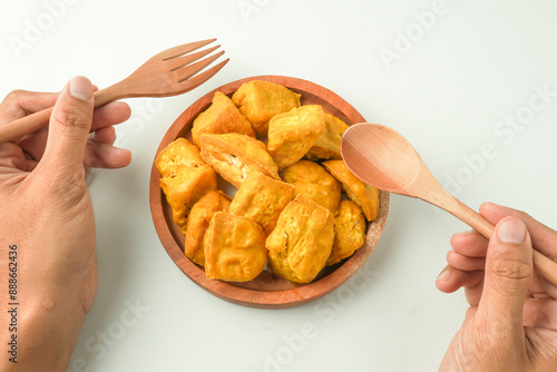 Someone hand holding wooden spoon and fork to eating yellow tofu on wooden plate. Tahu Takwa is a specialty food from Kediri, East Java, Indonesia. Flat Lay Photo photo