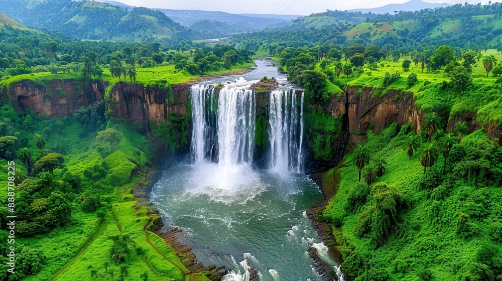Naklejka premium Breathtaking aerial view of a stunning waterfall cascading into a lush green valley surrounded by mountains and vibrant greenery.