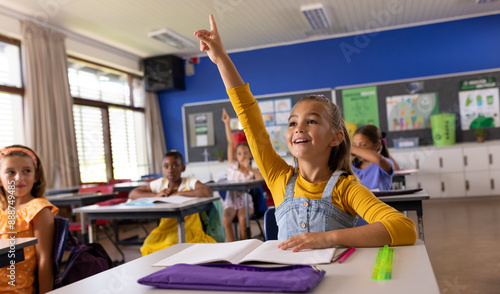 Caucasian schoolgirl raising hand while studying with diverse children in classroom