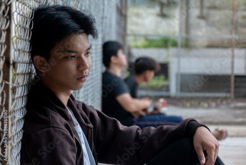 Schilderij op canvas Asian preteen boy in a black hoodie sits with his knees pressed against a metal fence panel in a juvenile detention facility, awaiting further release, freedom and detention of people concept
