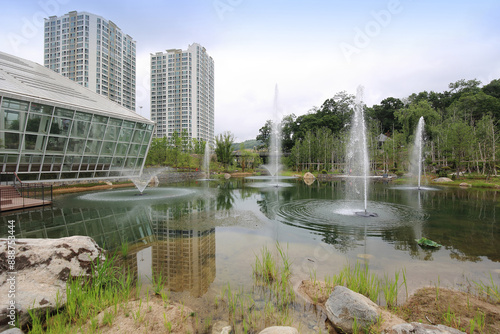 Yeongtong-gu, Suwon-si, Gyeonggi-do, South Korea - May 21, 2023: Summer view of water plant and fountain on the pond against greenhouse building and trees at Yeongheung Arboretum
 photo