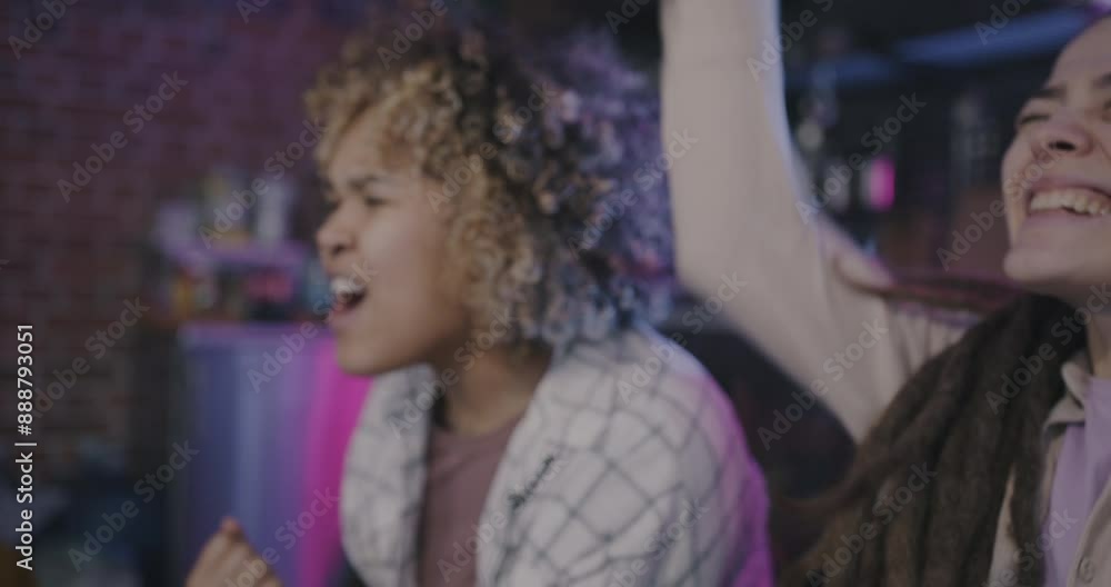 Two young women Caucasian and African American watching sports on TV cheering expressing joy at home in evening. Fans and hobby concept. Two young women Caucasian and African American watching sports on TV cheering expressing joy at home in evening. Fans and hobby concept.