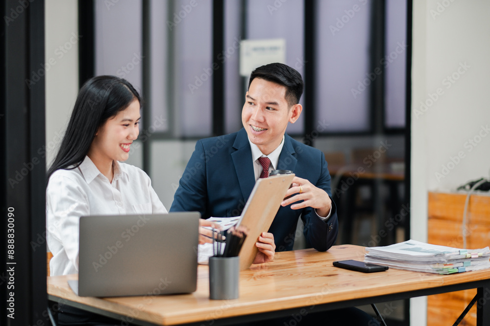 Two business professionals in a modern office setting, engaged in a discussion while reviewing documents and working on a laptop. Two business professionals in a modern office setting, engaged in a discussion while reviewing documents and working on a laptop.