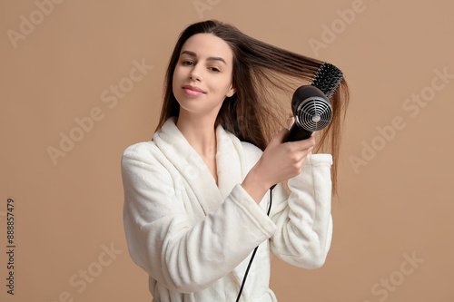 Young woman in bathrobe blow-drying hair with hairdryer on beige background photo