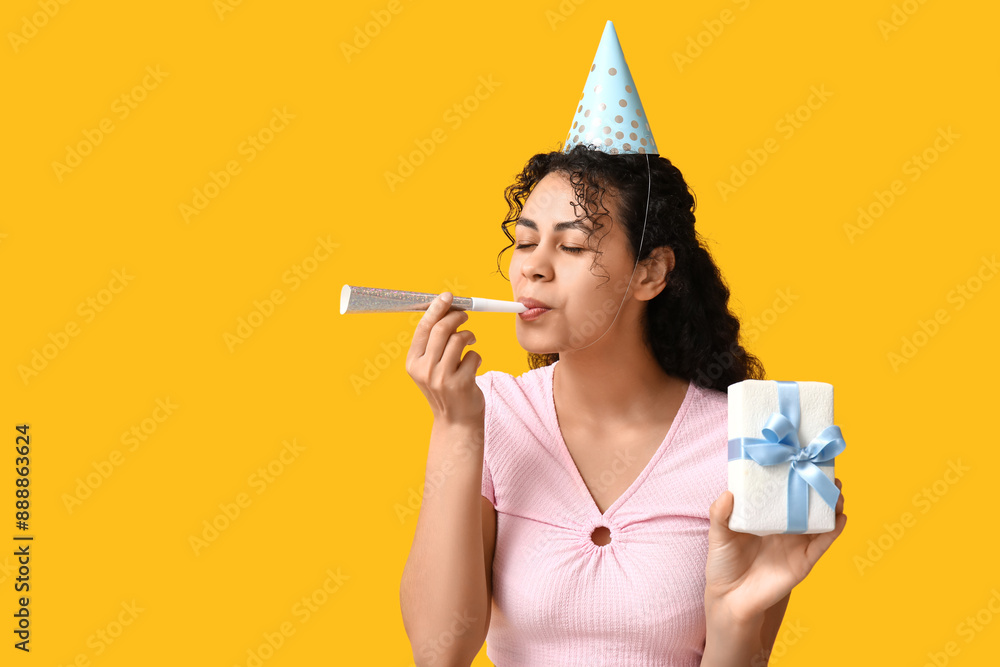 Beautiful young African-American woman in party hat with whistle and gift box on yellow background Beautiful young African-American woman in party hat with whistle and gift box on yellow background