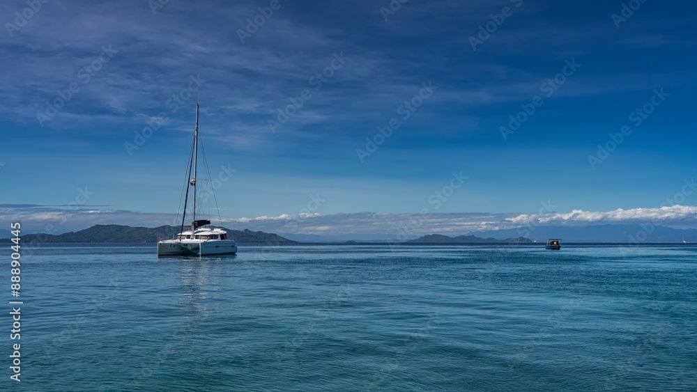 A beautiful white yacht is anchored in the ocean. A tall mast against a background of blue sky and clouds. Reflection on calm water. Mountains in the distance. Madagascar. Copy Space A beautiful white yacht is anchored in the ocean. A tall mast against a background of blue sky and clouds. Reflection on calm water. Mountains in the distance. Madagascar. Copy Space