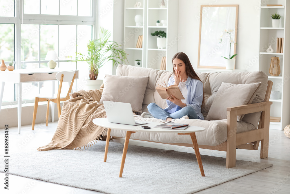 Beautiful young shocked woman with laptop reading book on sofa at home Beautiful young shocked woman with laptop reading book on sofa at home