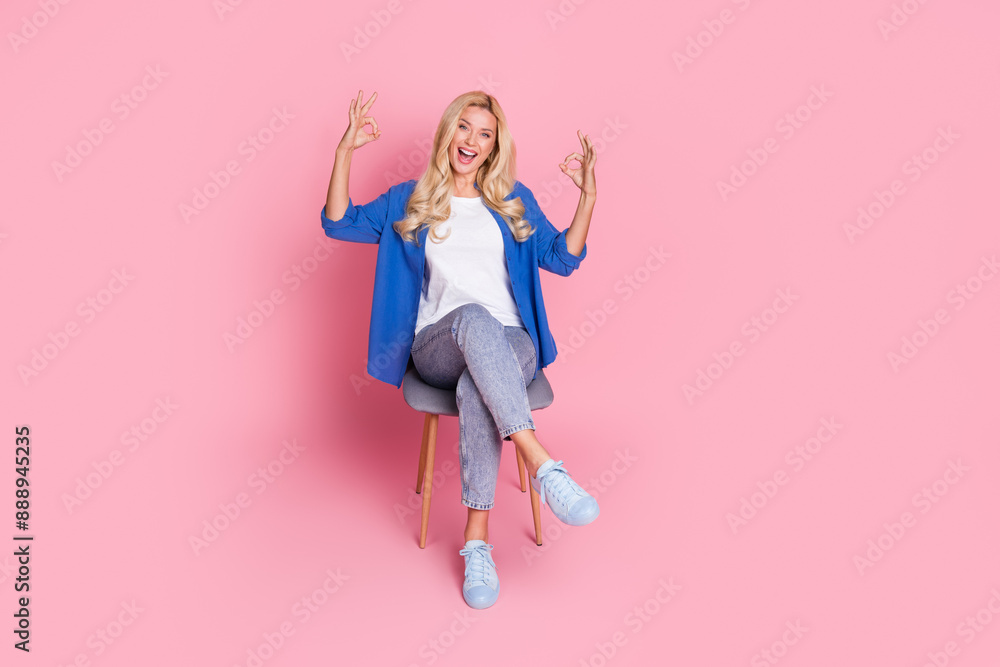 Full size photo of adorable cheerful woman dressed blue shirt jeans sitting on chair showing okey isolated on pink color background Full size photo of adorable cheerful woman dressed blue shirt jeans sitting on chair showing okey isolated on pink color background