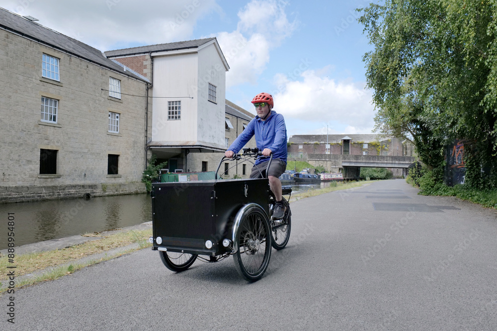 Obraz premium White middle aged man rides a large black electric cargo bike along a canal towpath in the summer.