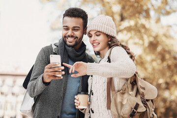 Beautiful happy young couple using smartphone outdoors. Joyful smiling woman and man looking at mobile phone in a city, autumn portrait. Love, technology, communication, winter holiday travel concept