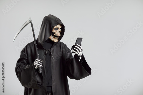 Konstfotografi Awful grim reaper, with a scythe and wearing skeleton mask, uses a mobile phone on a grey background