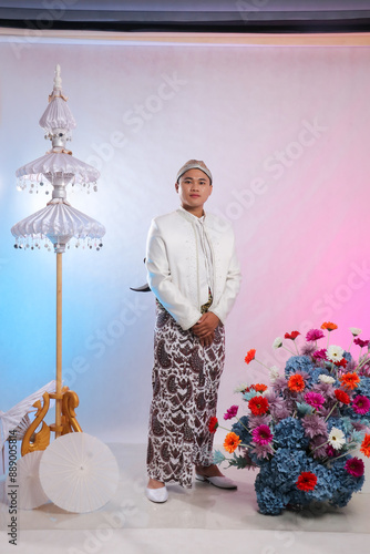 Obraz na płótnie full body gallantry of young elegant Asian man standing among flower decorations wearing modern Javanese traditional clothing, white velvet bridal kebaya on white background