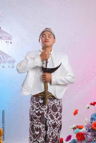 Foto The gallantry of an elegant young Asian boy standing taking a keris up wearing modern Javanese traditional clothing, white velvet bridal kebaya on a white studio background