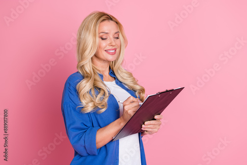 Portrait of friendly adorable woman with wavy hair dressed blue shirt signing documents on clipboard isolated on pink color background