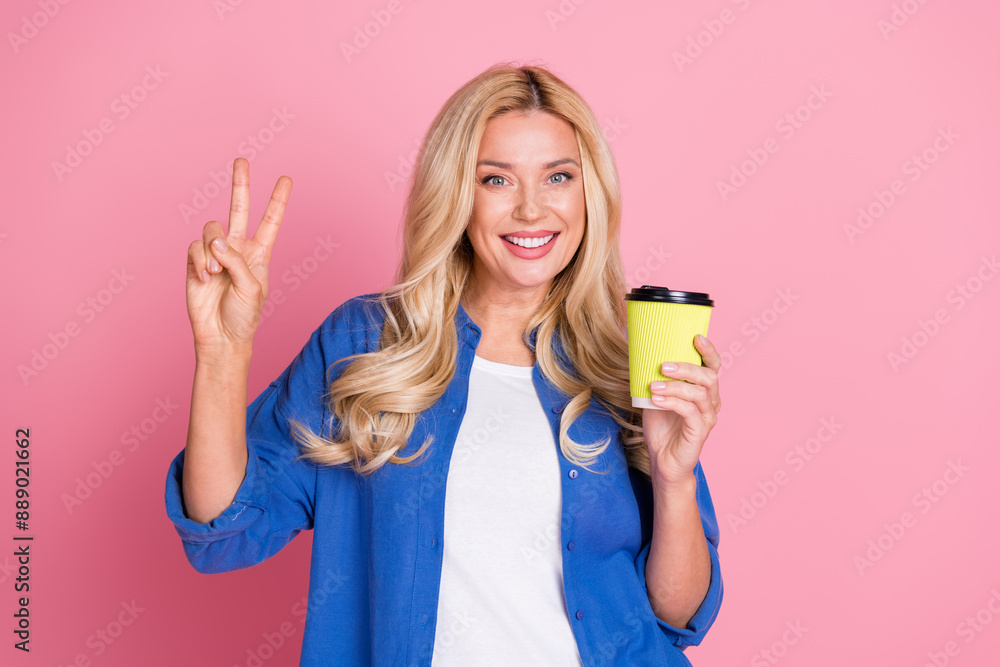 Photo of optimistic woman with wavy hairstyle dressed blue shirt hold cup of coffee showing v-sign isolated on pink color background Photo of optimistic woman with wavy hairstyle dressed blue shirt hold cup of coffee showing v-sign isolated on pink color background