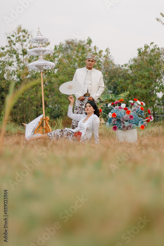 couple young indonesia male and female lovers smiling, women holding umbrellas sitting under modern Javanese traditional clothing, bridal white velvet kebaya in an outdoor barren garden photo