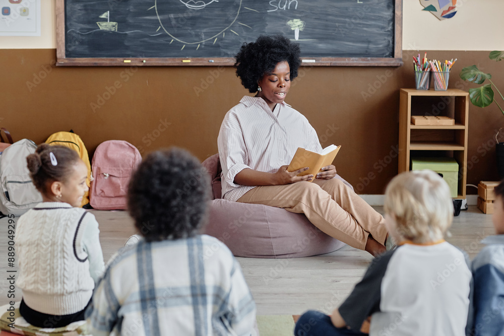 © Seventyfour - Thoughtful female teacher of Black ethnicity in bean bag chair reading book aloud to primary school students sitting around on classroom floor © Seventyfour - Thoughtful female teacher of Black ethnicity in bean bag chair reading book aloud to primary school students sitting around on classroom floor