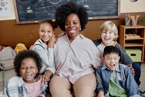 Portrait shot of smiling female teacher of Black ethnicity sitting in chair near multiethnic happy children hugging her in classroom photo