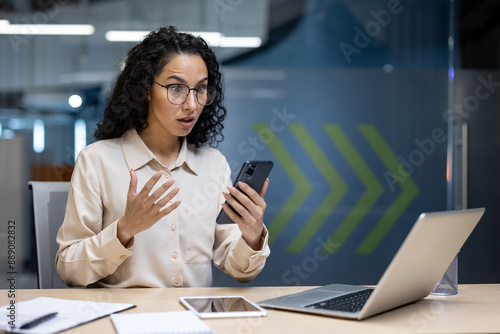 Shocked businesswoman looking at smartphone in office. Woman reacting to surprising or alarming news. Workspace with laptop, tablet, and notepad. Indoor professional setting.