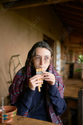 A young woman is sitting at a rustic wooden table playing a wooden pan flute. They are wearing eyeglasses, a plaid shawl, and have a contemplative expression. photo