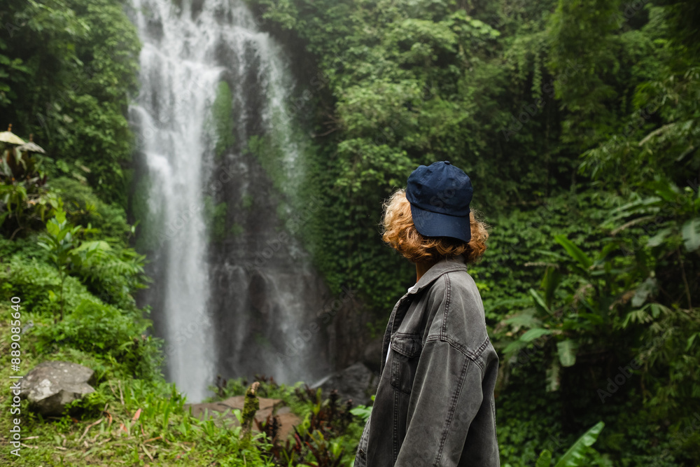 A young girl, a traveler, admires a stunning waterfall in the jungle. Back view. A young girl, a traveler, admires a stunning waterfall in the jungle. Back view.