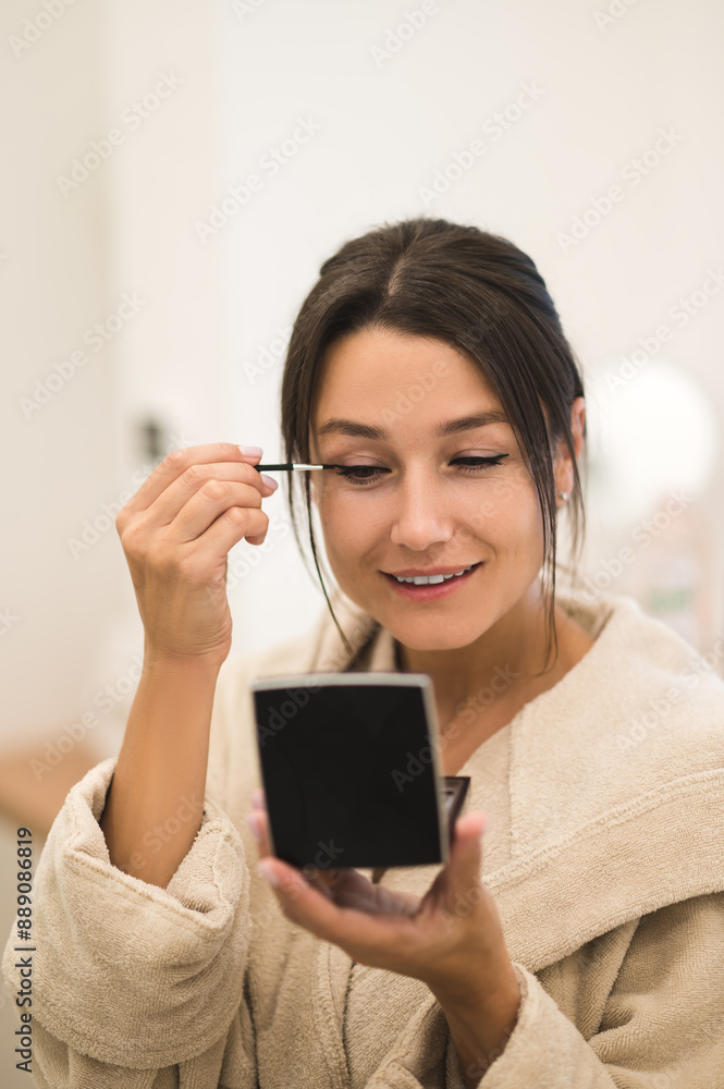 Cute dark-haired young woman doing her make up and looking contented Cute dark-haired young woman doing her make up and looking contented