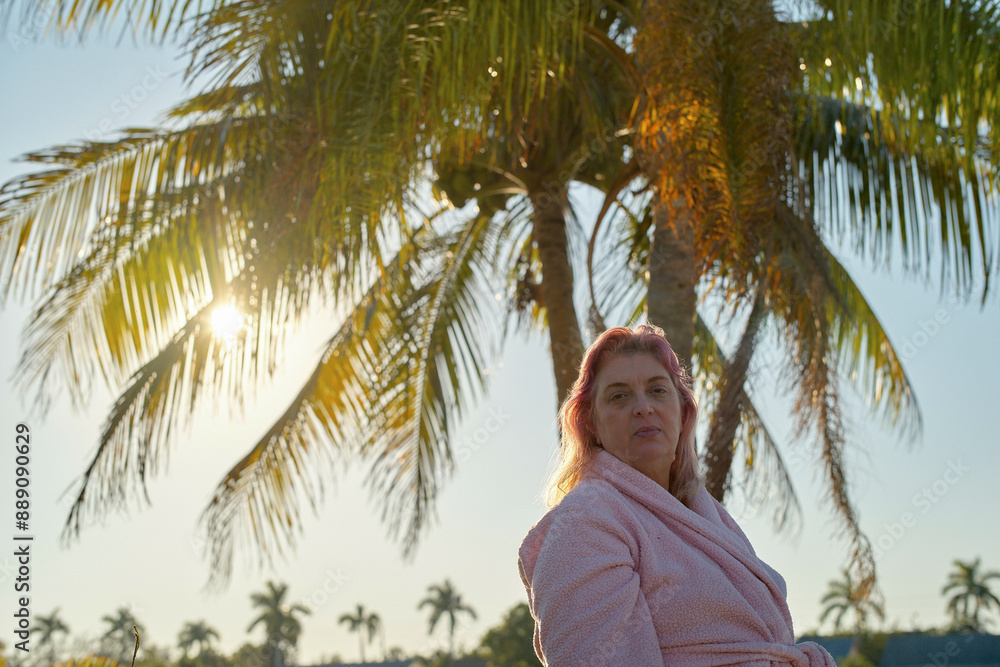 Woman in a pink bathrobe stands in front of palm trees with the sun setting in the background. Woman in a pink bathrobe stands in front of palm trees with the sun setting in the background.