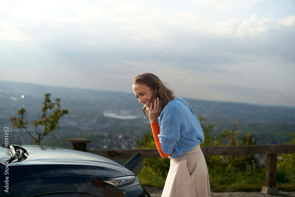 Smiling woman standing by a car overlooks a cityscape from a high vantage point at sunset Smiling woman standing by a car overlooks a cityscape from a high vantage point at sunset