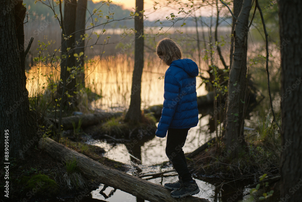 A young child in a blue jacket stands on a log by a tranquil forest lake at sunset. A young child in a blue jacket stands on a log by a tranquil forest lake at sunset.