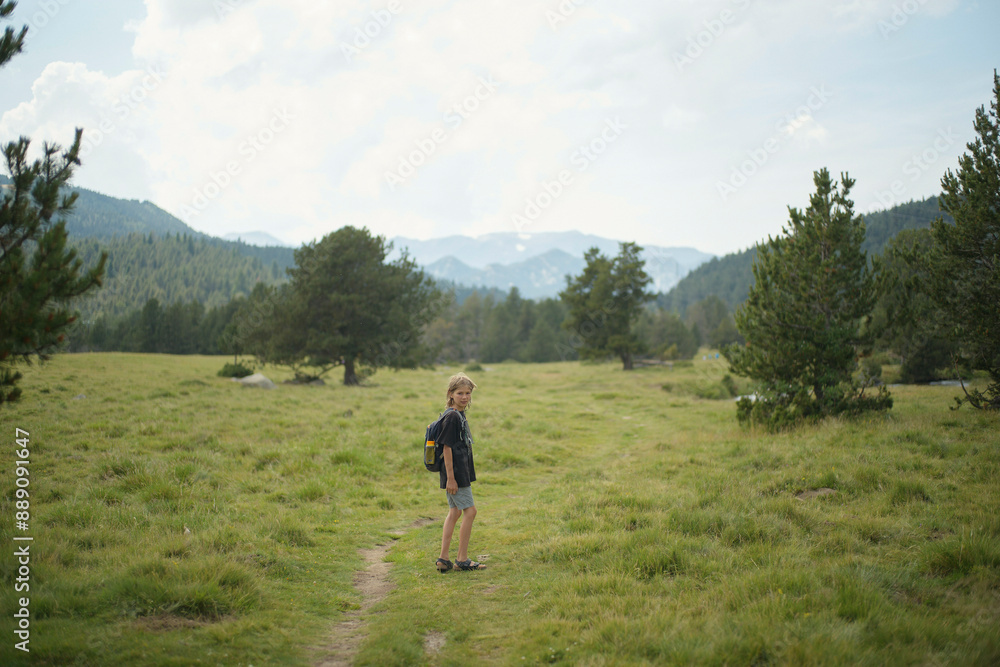 A child with a backpack stands on a grassy trail in a scenic mountain landscape, surrounded by trees under a cloudy sky. A child with a backpack stands on a grassy trail in a scenic mountain landscape, surrounded by trees under a cloudy sky.