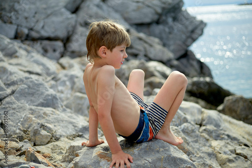 A young child in striped swimwear sitting on rocks by the sea, looking over their shoulder with a joyful expression.