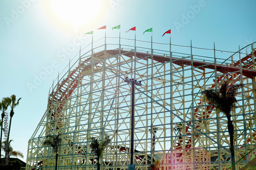 Sunlight flares over a towering wooden roller coaster adorned with colorful flags under a clear blue sky. photo