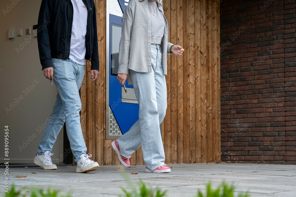 Two people casually walking past a modern building with a wooden facade and brick wall, captured from the waist down. Two people casually walking past a modern building with a wooden facade and brick wall, captured from the waist down.