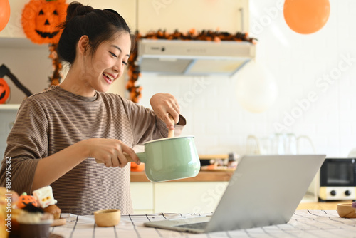 фотографія Happy young woman cooking and following an online recipe on laptop in a kitchen