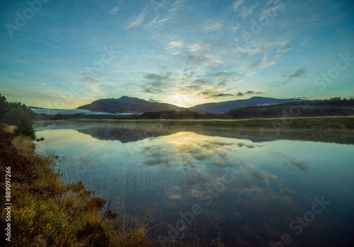Sunrise over a tranquil lake with reflections of clouds and hills on the water surrounded by forested landscape.