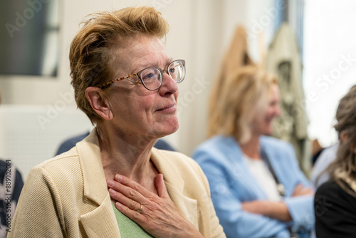 A woman with glasses wearing a beige jacket is touching her chest with a hand, showing an expression of gratitude or relief. photo