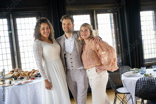 Smiling bridal couple with a guest posing in a banquet hall with a table filled with food.